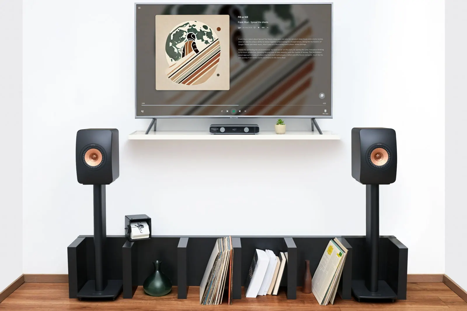Un salón con un televisor de pared que muestra la carátula de un álbum, dos altavoces sobre pies y Primo Plus en una estantería baja con discos.