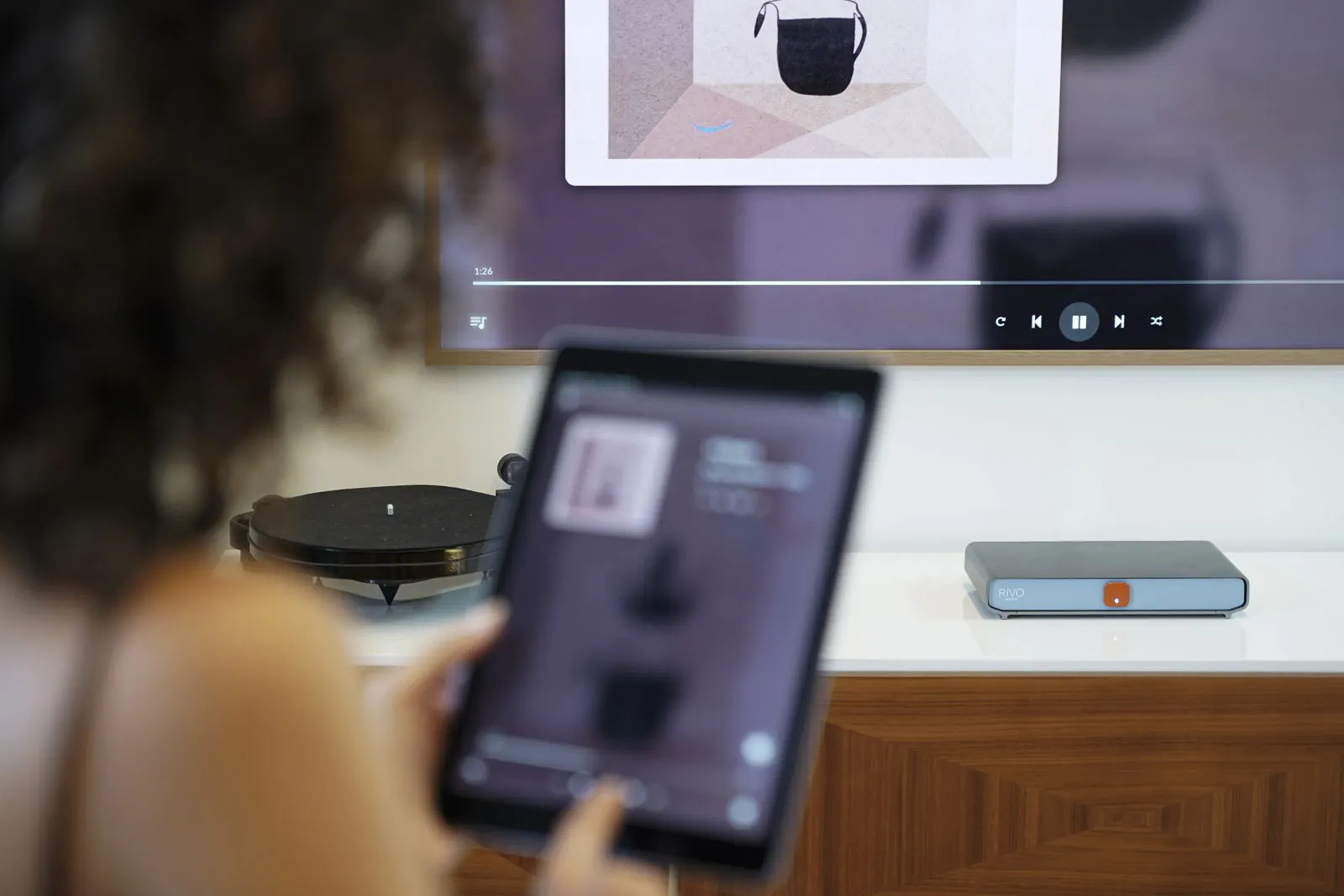 Un ascoltatore tiene in mano un tablet che mostra la copertina di un album, con Rivo su una credenza e lo stesso album sulla TV alle sue spalle.