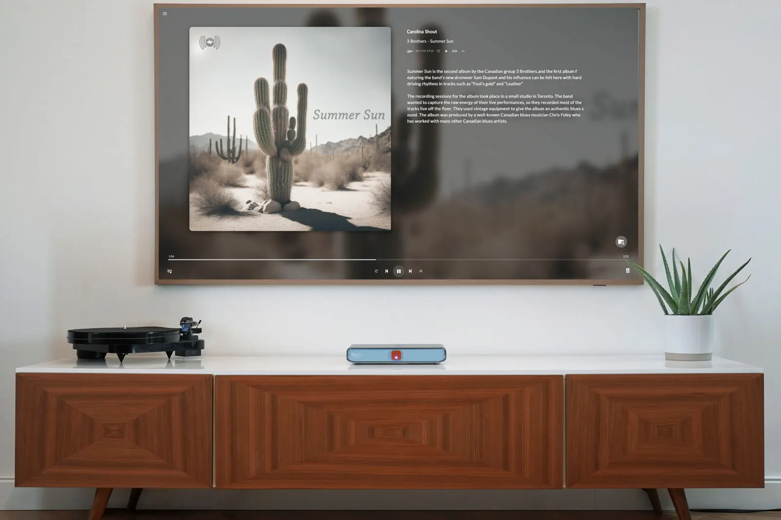 Un soggiorno con una TV a parete che mostra la copertina di un album con un cactus e i dettagli del brano, sopra una credenza di legno con un giradischi e Rivo.
