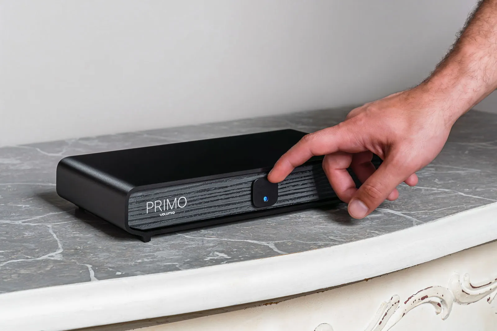 Una mano che raggiunge il pulsante sul frontale di Primo V3, appoggiato su un piano di marmo grigio.