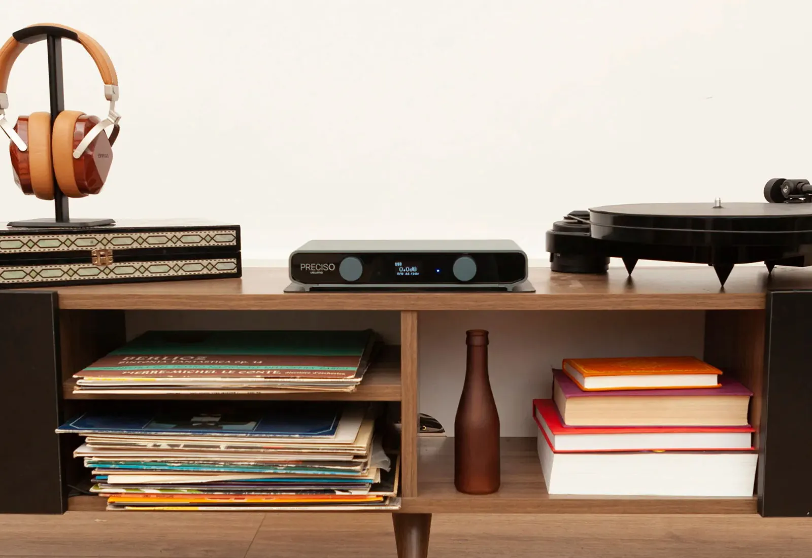 Preciso su una credenza di legno tra un paio di cuffie su un supporto e un giradischi, con dischi, libri e una bottiglia marrone sui ripiani sottostanti.