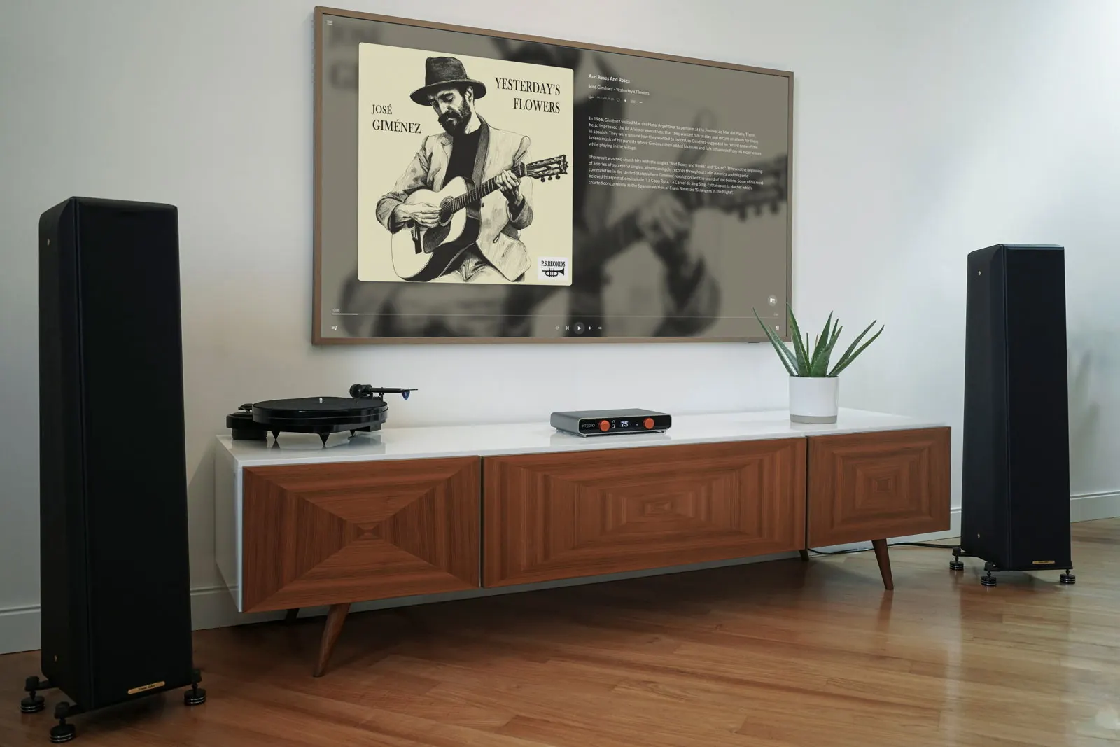 Ein Wohnzimmer mit Integro auf einem Holz-Sideboard zwischen zwei hohen schwarzen Standlautsprechern, neben einem Plattenspieler und einer Pflanze, unter einem Wandfernseher, der ein Albumcover zeigt.