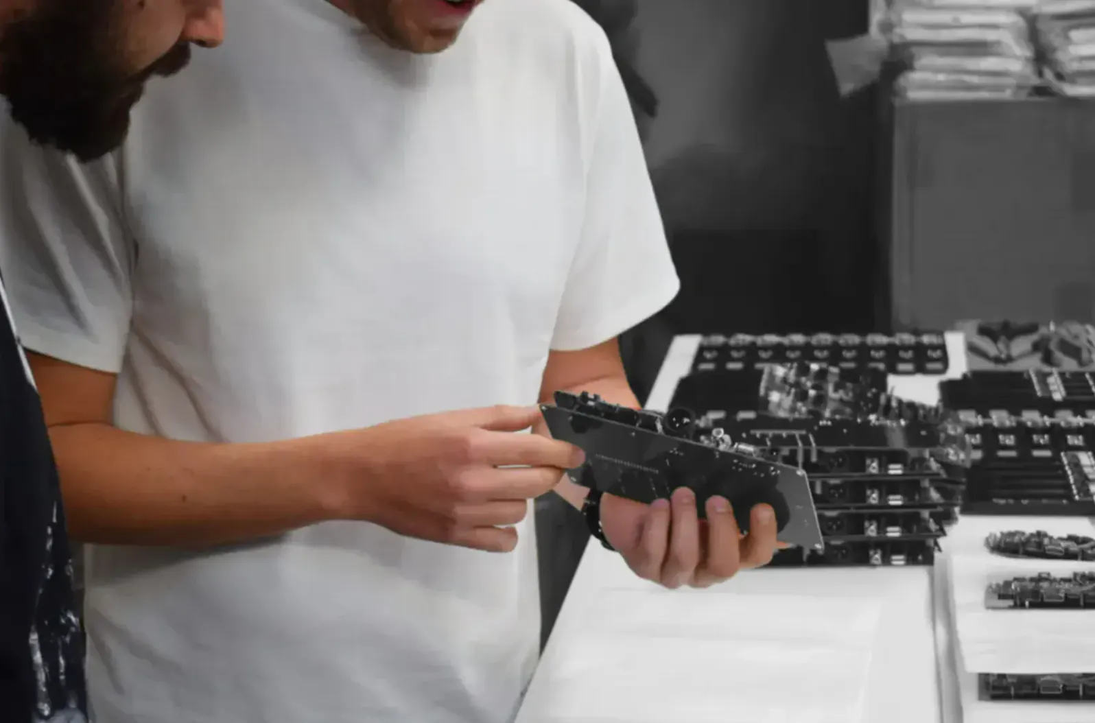 An einer Werkbank hält eine Person in weißem T-Shirt eine dunkle Platine über Reihen weiterer Platinen.