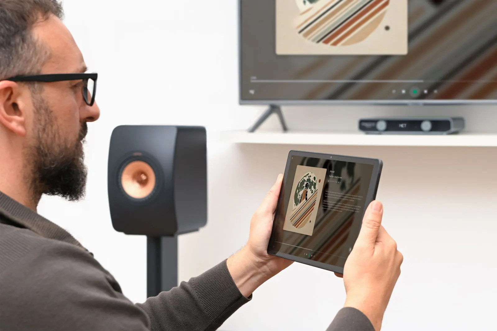 Una persona sostiene una tablet con la app de Volumio, junto a un altavoz y un televisor.