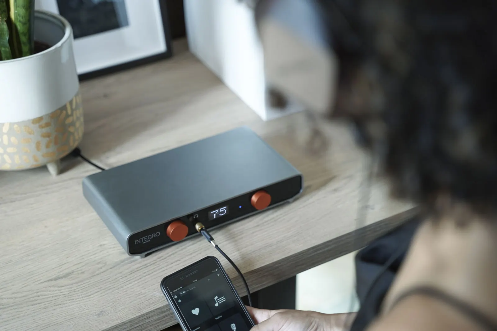 Ein Hörer hält ein Smartphone mit einer Musikbibliothek, neben Integro auf einem Holztisch, vorne sind Kopfhörer eingesteckt.
