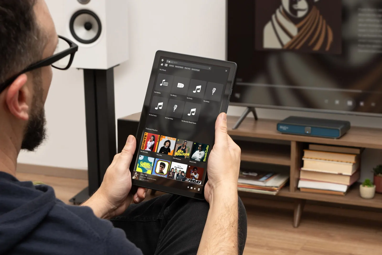 Ein Hörer hält ein Tablet mit einer Musikbibliothek, hinter ihm Rivo Plus auf einem Sideboard unter dem Fernseher.