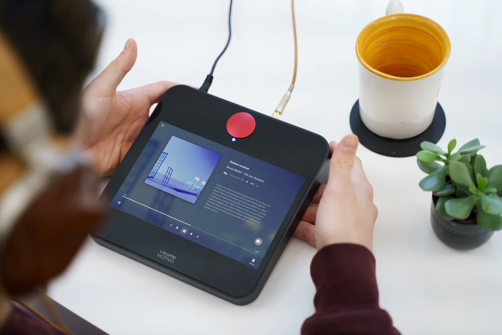 Two hands holding Motivo on a white table, with its touch screen showing an album and headphone and power cables plugged in at the back.